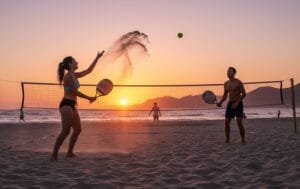 Dupla jogando beach tennis na areia, com raquete e bola em destaque sob o sol.
