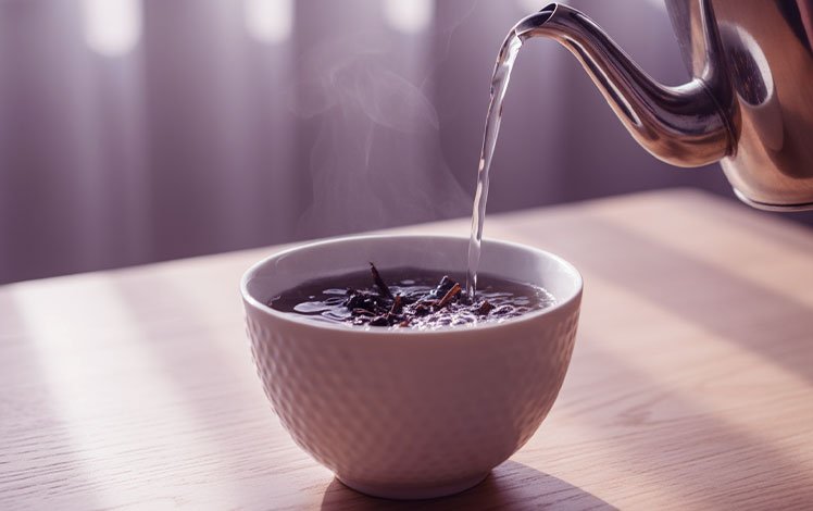 Água quente sendo despejada sobre folhas de chá em xícara branca, com vapor subindo e luz suave, representando calma e presença.