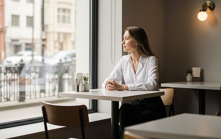 mulher sozinha em café, olhando para fora, representando reflexão e solidão emocional.