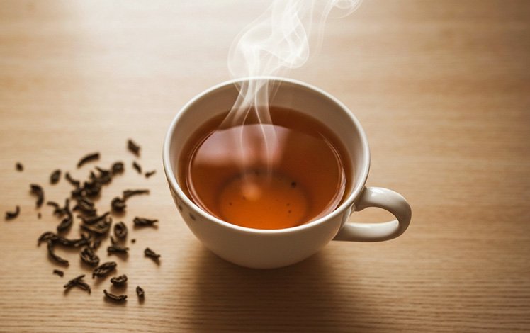 Xícara de chá quente vista de cima com folhas de chá ao redor sobre mesa de madeira clara